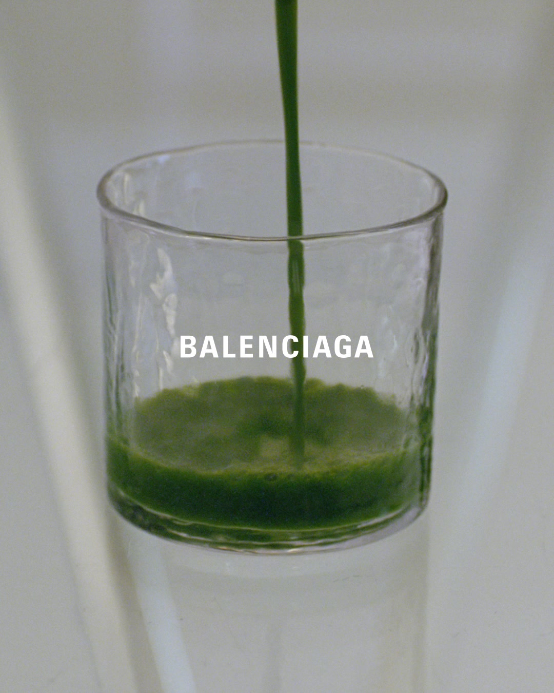 A close-up shot shows green liquid being poured into a clear, textured glass. The Balenciaga logo is overlaid in white text across the middle of the glass. The background is softly blurred and light-colored, creating a minimalist aesthetic. The image focuses on the act of pouring and the branding.