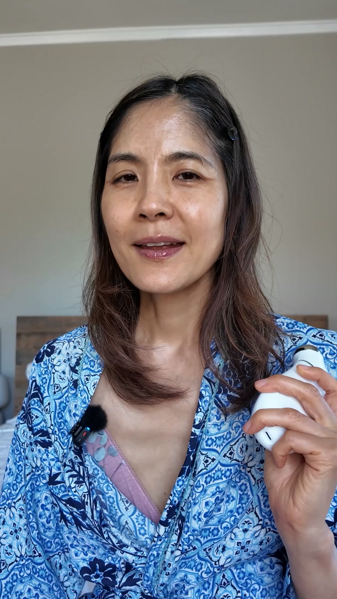 A woman with medium-length brown hair, wearing a blue and white patterned robe, holds a white NuFace device in her right hand. She is looking directly at the camera with a slight smile. Text overlays the video, stating the product's benefits and name. The background appears to be a bedroom with a bed and headboard visible. A small black microphone is clipped to her robe.