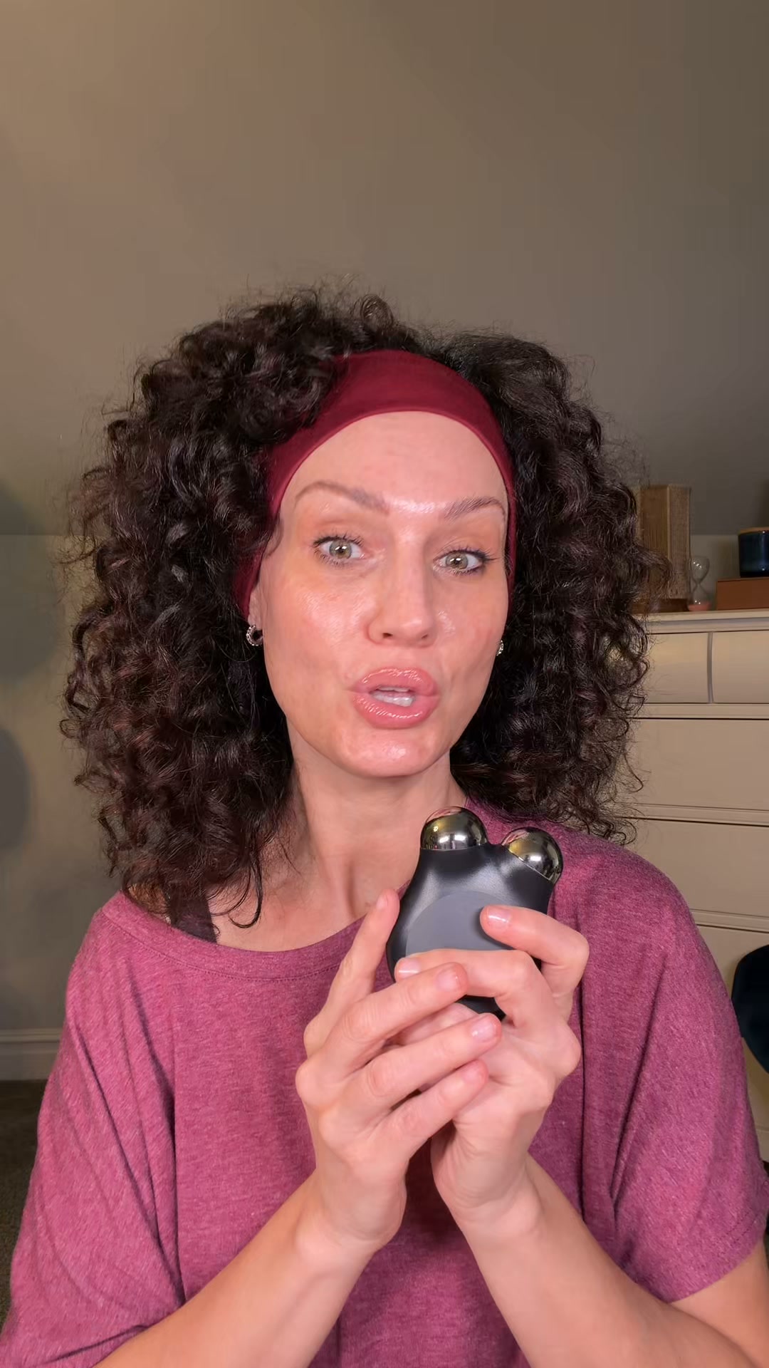 A woman with curly dark hair, wearing a maroon headband and a maroon t-shirt, holds a black NuFace device with two silver spheres in her hands. She looks directly at the camera with a slight smile and parted lips. The background is a plain, neutral wall with a dresser and some decorative items visible on the right side. Text overlays the image, highlighting a special offer: 'Better Together ⚡ Buy the NUFACE MINI+ and get a FREE FIX Microwand. Two skincare essentials, one limited-time offer. Shop now!'