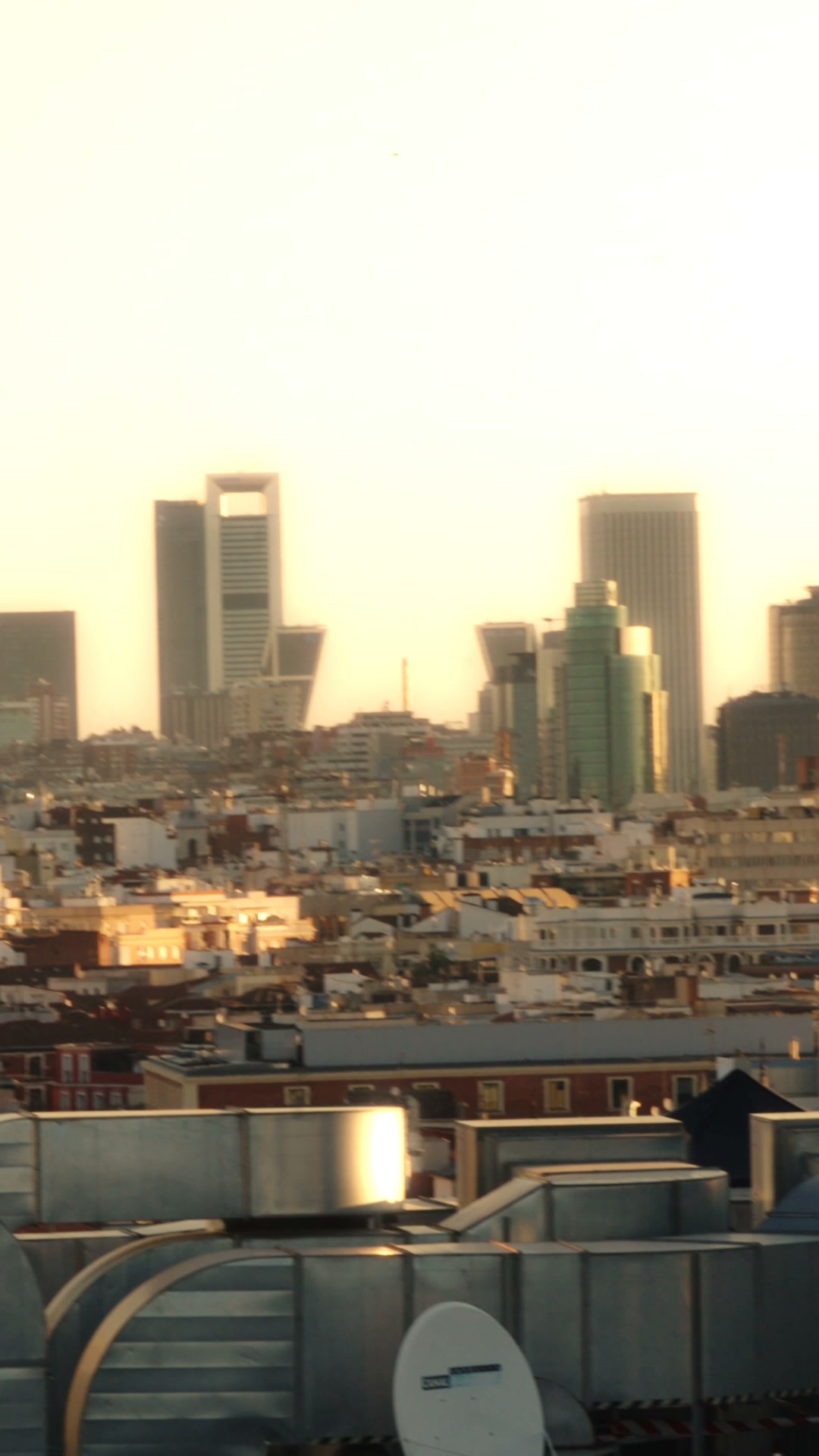 A panoramic view of a city skyline at sunset. The sky is a hazy yellow, and the buildings are silhouetted against it. In the foreground, there are several large, metallic air conditioning units and a satellite dish. The text overlay reads "Everyday Seamless, now in new colours 💛🩷 Come try the soft, second-skin gym sets you'll want to wear every day."