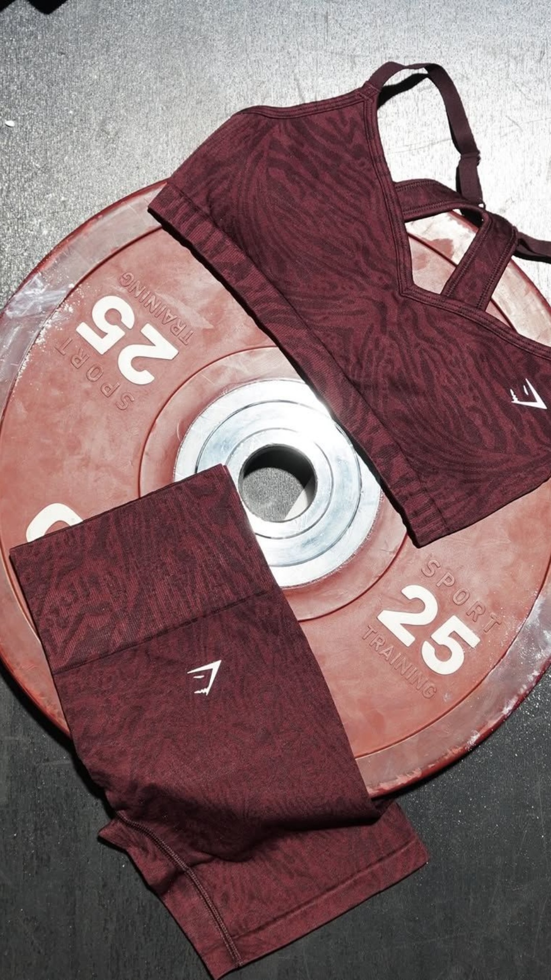 A flat lay image shows a burgundy workout set with a subtle animal print pattern. The set, consisting of a sports bra and leggings, is placed on top of a red 25lb weight plate. The Gymshark logo is visible on the leggings. The text overlay wishes the viewer a great year and offers a discount code for their first order.
