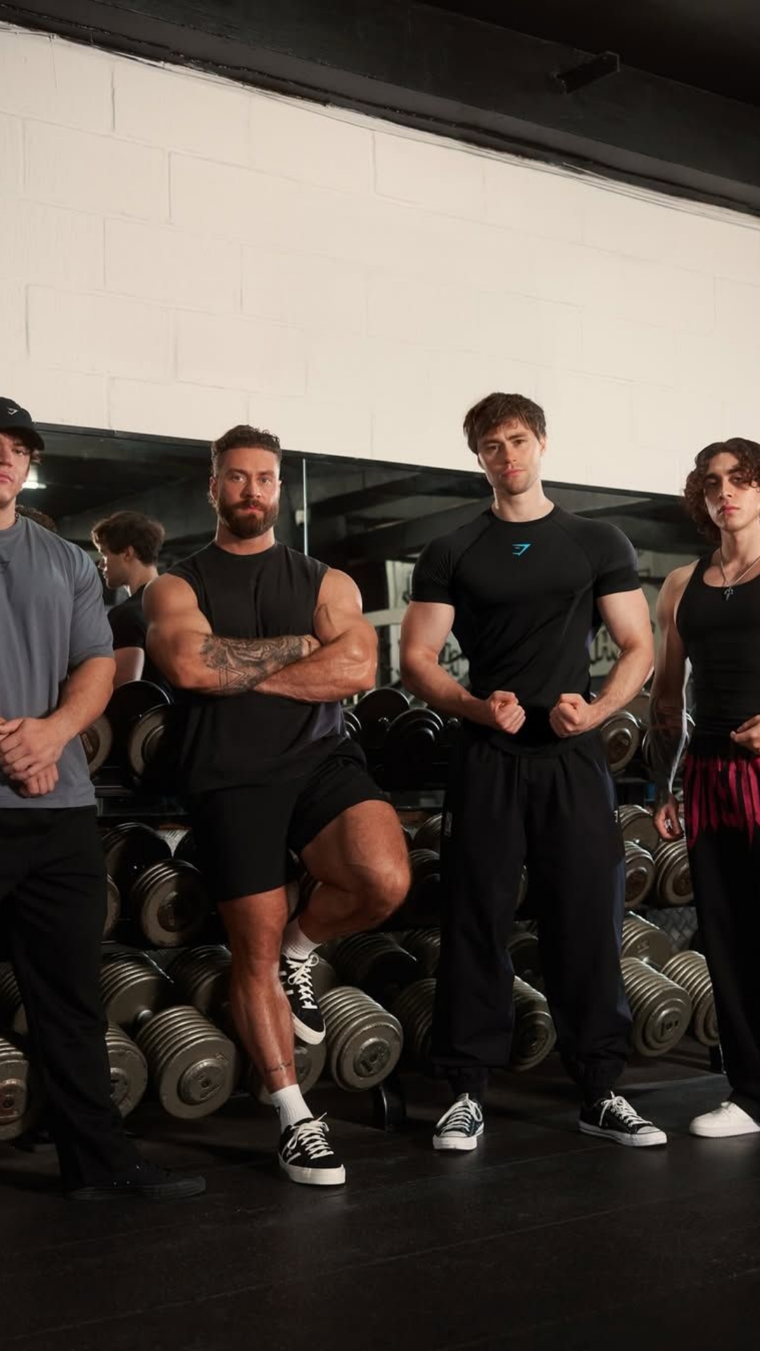 Four men in athletic wear stand in a gym in front of a rack of dumbbells. The man in the center is muscular with a beard and is posing with his arms crossed. The other men are also posing, with two flexing their biceps. The Gymshark logo is visible on the shirt of one of the men. Text overlayed on the image reads: "The gym isn’t just what we do best, it’s everything we do🏋️‍♂️ We Do Gym🦈".