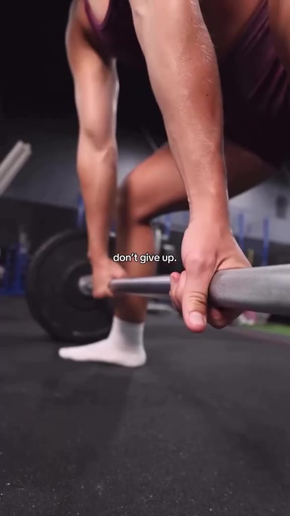 A close-up shot focuses on a person's hands gripping a barbell during a weightlifting exercise. The individual is wearing white socks and dark athletic shorts. Text overlaid on the image reads "don't give up." The background is a gym setting with blurred equipment and blue accents.