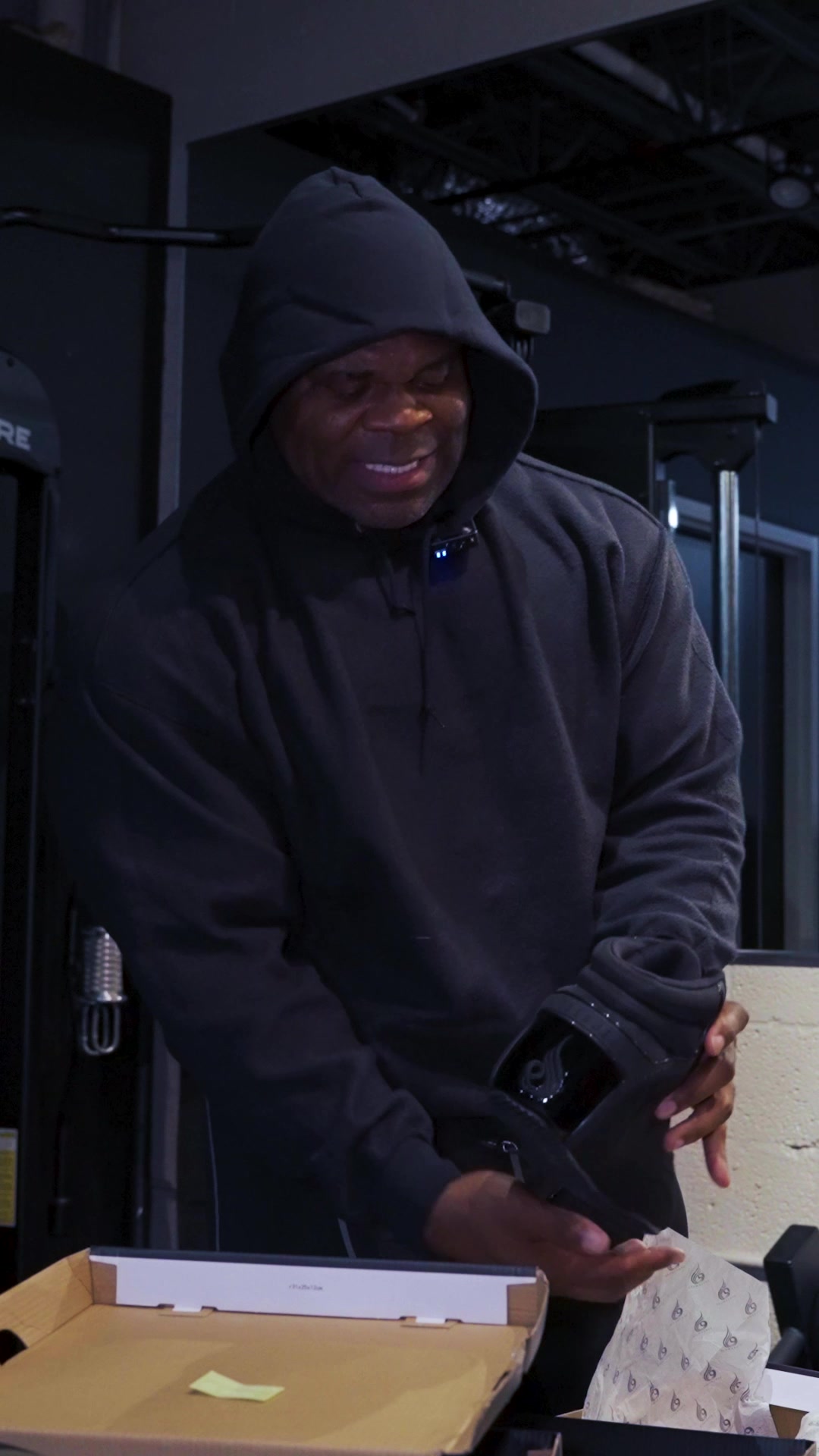 A man wearing a black hoodie stands in a gym setting, holding a black shoe. He is looking down at the shoe with a smile. In front of him is an open box with tissue paper. The shoe is black with a shiny logo on the side. Text overlays the image, introducing the D-Mak Bodybuilding Shoes and highlighting their features for optimal power, stability, and drive, recommending them for seasoned lifters.