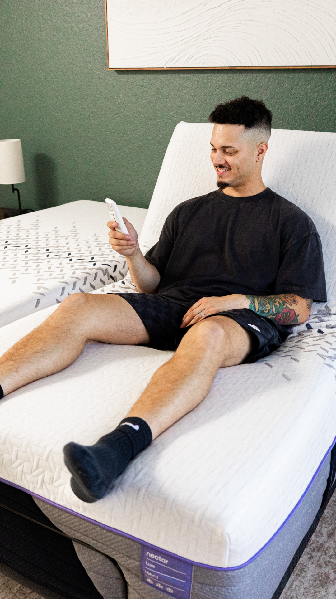 A man is sitting on an adjustable bed base with a Nectar Luxe Hybrid Split King mattress, holding a remote control and smiling. He is wearing a dark t-shirt and patterned shorts. The bed is in a bedroom with a green textured wall and a framed piece of art above the bed. Text overlays highlight the adjustable base features like zero gravity, anti-snore, massage mode, and USB ports. The copy asks if the viewer has invested in their sleep and details the benefits of the Nectar adjustable base and mattress bundle.