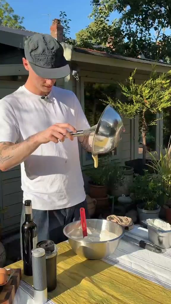 A man wearing a white t-shirt and a black cap is outdoors, preparing food in a large metal bowl. He is holding a whisk and pouring a yellow liquid from a metal bowl into the larger bowl. On the table are a bottle of wine, a thermos, eggs, and other ingredients. The background shows a garden setting with trees and a small building. The text overlay reads: "Parfois, on a juste envie de sortir de la cuisine et de cuisiner dans le jardin."