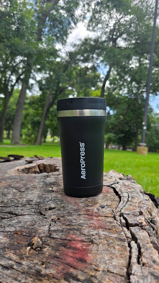 A black AeroPress Go Plus travel coffee maker is placed on a weathered, cracked tree stump in an outdoor setting with green grass and trees in the background. The product has the "AeroPress" logo printed vertically on its side. The text overlay reads: "There’s something different about that first cup of coffee before the day begins. Going for a walk and brewing on the tailgate of my truck just hits different this summer. The AeroPress Go Plus made it easy to pack everything I needed into one compact travel system, brew a smooth cup in just 2-3 minutes, and get moving without slowing down. Its 3-in-1 brewing technology delivers a rich, grit-free cup wherever your adventures take you. If you're planning road trips, camping weekends, or just want better coffee on the go, now's the perfect time to grab one. Use my code coffeewitheto to save on your order!"