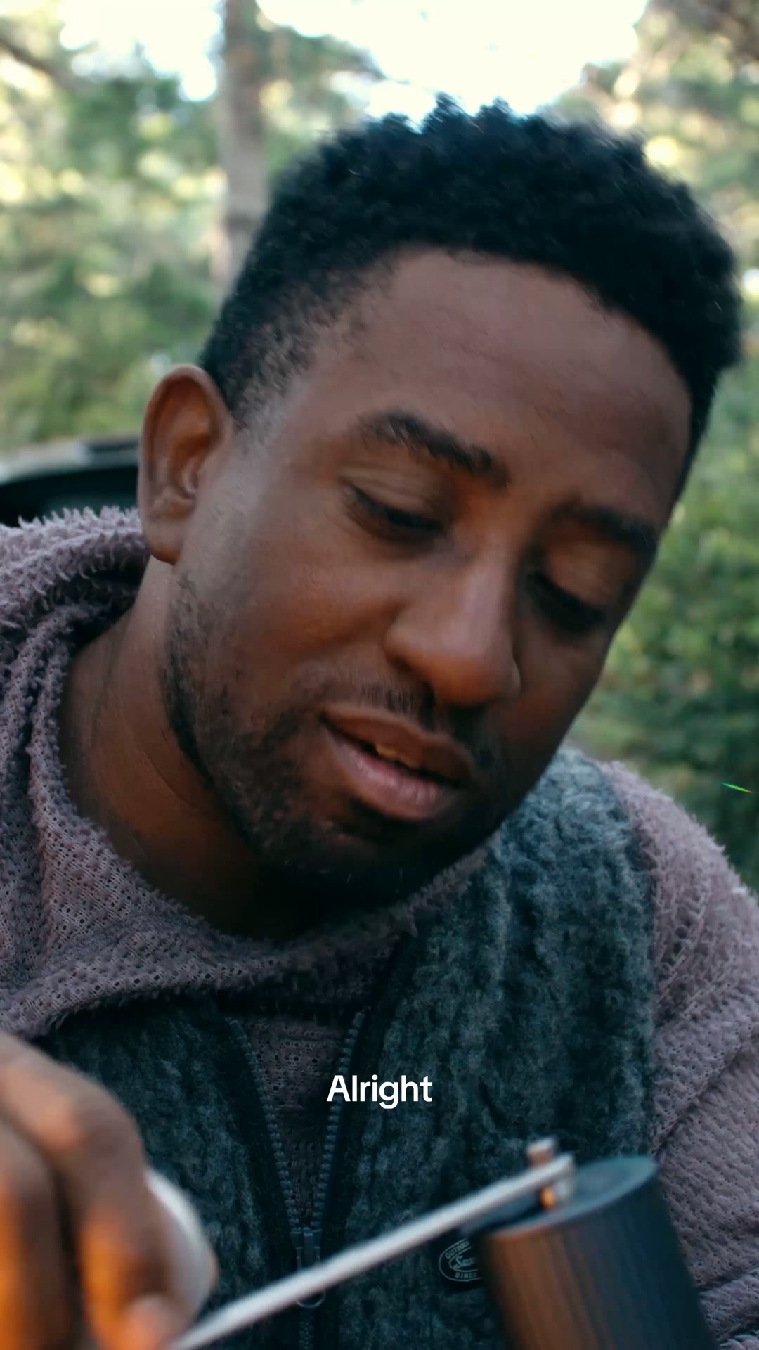 A close-up shot of a Black man with short, curly hair, wearing a grey fleece vest over a purple hoodie. He is looking down and to the right, with a slight smile. The background is blurred, showing trees and foliage, suggesting an outdoor setting. White text overlaid on the image reads "Alright".