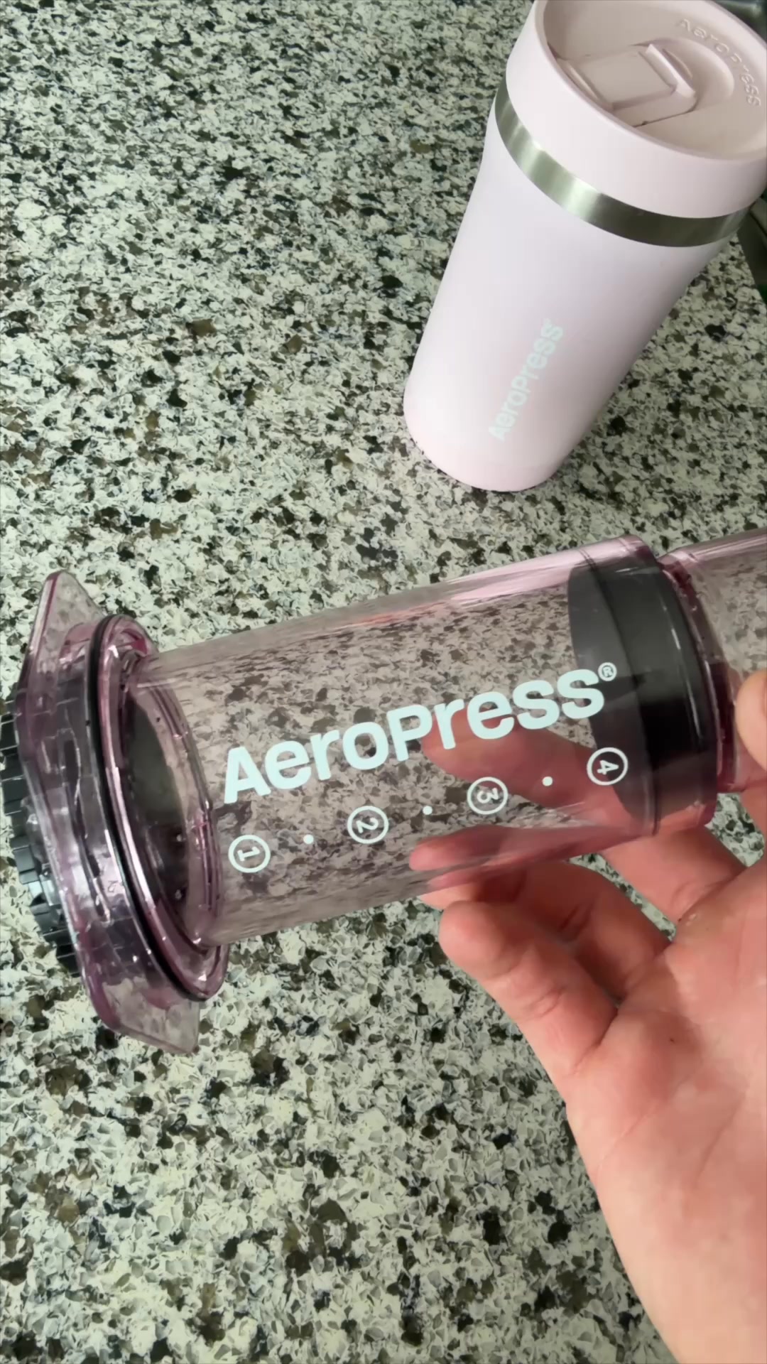 A close-up shot shows a person holding a clear, purple AeroPress coffee maker. In the background, a pink AeroPress travel mug sits on a speckled countertop. White text overlays the coffee maker, featuring a quote about the quality of coffee made with the product. Additional text highlights key features like smooth flavor, patented technology, and quick brewing and cleaning.