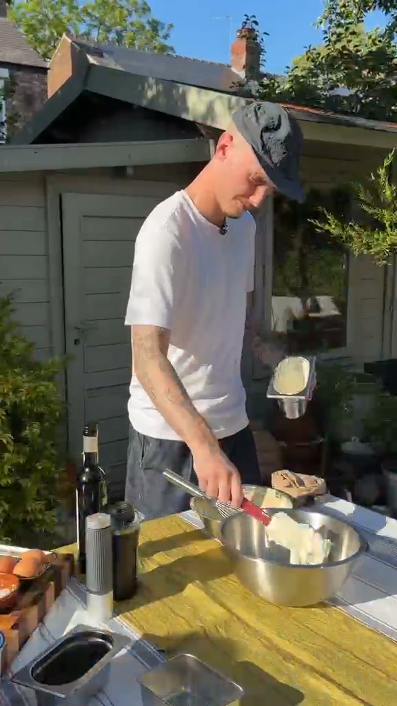 A man wearing a white t-shirt and a dark baseball cap is standing outside in a garden setting, preparing food in a metal bowl. He is holding a whisk and appears to be mixing ingredients. On the table in front of him are eggs, bottles, and other food preparation items. The text overlay reads: "A veces lo que te apetece es salir de la cocina y cocinar en el jardín, así que hoy te enseñamos lo fácil que es con la @aeropress: ¡el tiramisú de jardín!"