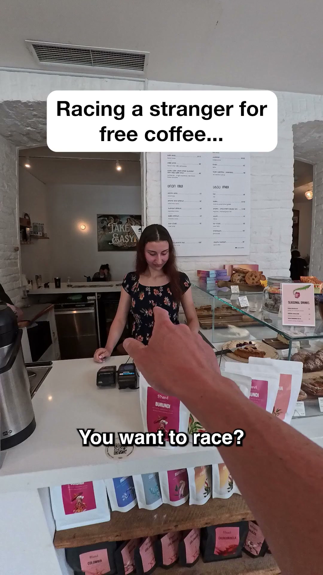 A first-person perspective shot inside a coffee shop. The camera is pointed towards a young woman behind the counter, who is smiling. Text overlays read "Racing a stranger for free coffee..." and "You want to race?". Bags of coffee beans are visible on the counter and shelves.