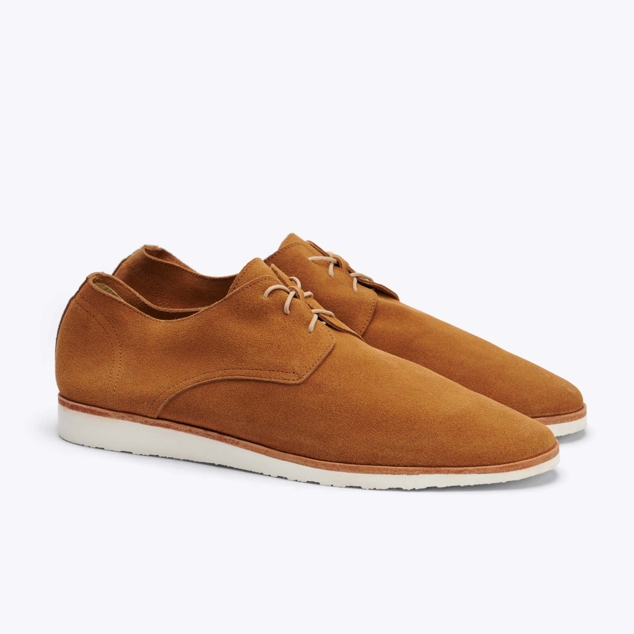 A pair of tan suede derby shoes with white soles is shown against a plain grey background. The shoes are angled to show the side profile and the lacing. The copy states that every purchase guarantees living wages in the factory in Peru, enables the pursuit of living wages throughout the supply chain, and offsets carbon emissions.