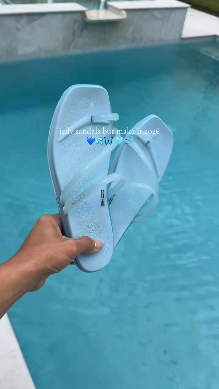 A hand holds up a pair of light blue Crocs jelly sandals over a bright blue swimming pool. Text overlaid on the image reads "jelly sandals but make it 2026" with blue butterfly and water drop emojis. The sandals appear to be a modern take on a classic style, suggesting a future trend. The overall aesthetic is summery and relaxed, fitting for a poolside setting.