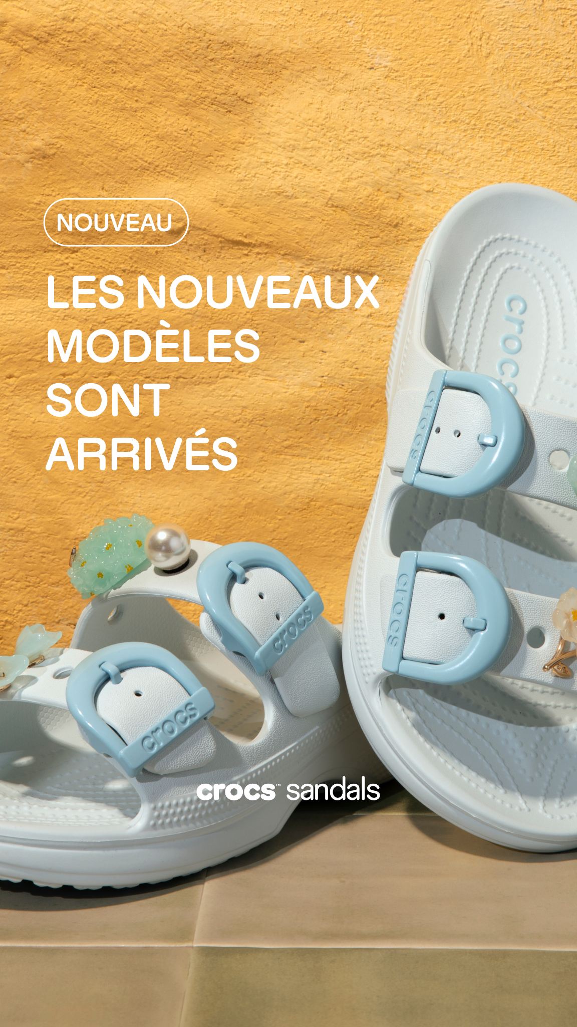 A close-up shot showcases two white Crocs sandals adorned with light blue buckles and decorative charms. The sandals are placed on a textured yellow surface, possibly sand, with a tiled surface below. White text overlays the image, announcing "NOUVEAU" in an oval shape and "LES NOUVEAUX MODÈLES SONT ARRIVÉS" in larger font. The Crocs logo and "sandals" are visible at the bottom right. The creative highlights new arrivals with a focus on personal expression.
