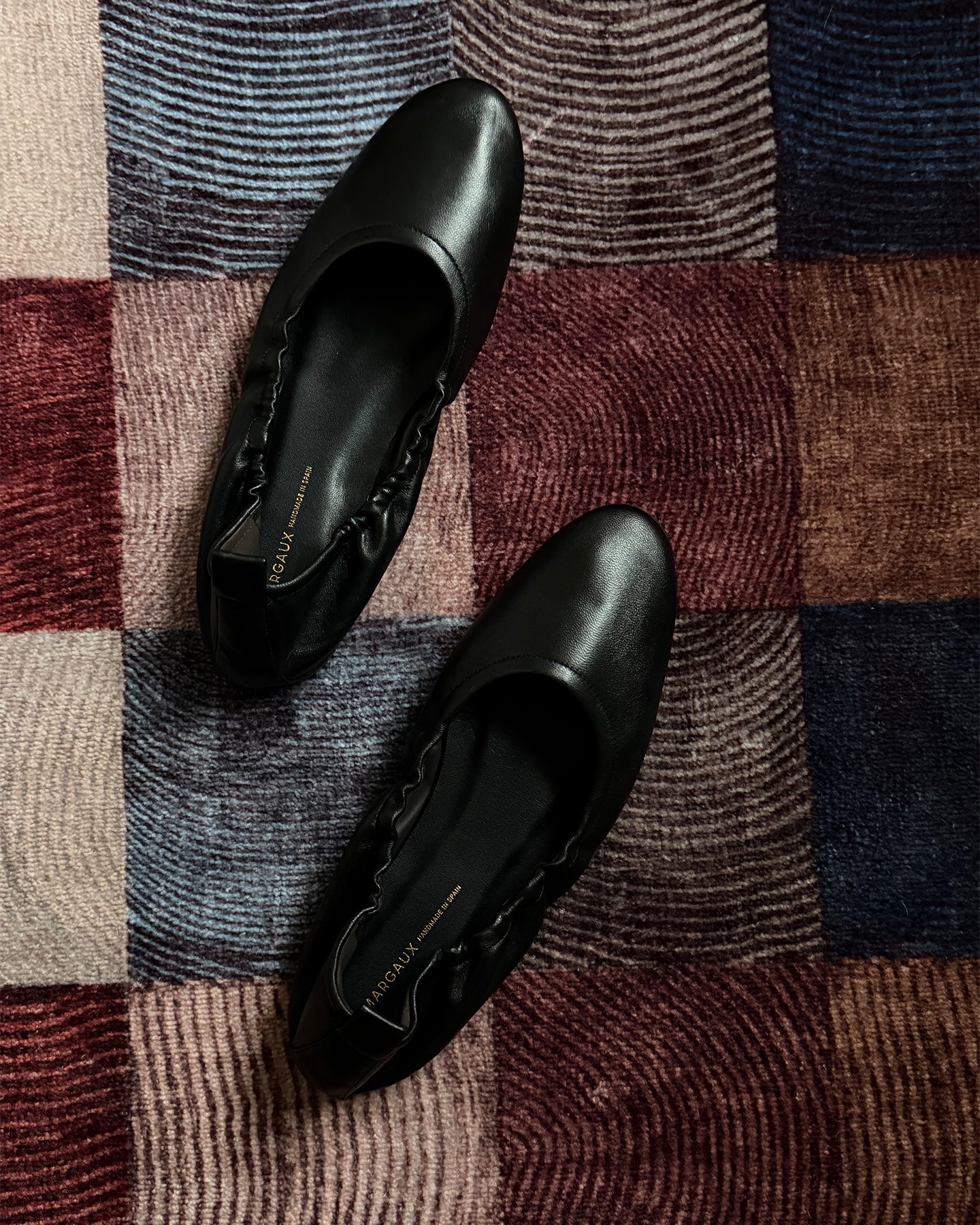A top-down, flatlay view of a pair of black leather ballet flats is shown on a textured, multi-colored rug. The shoes are positioned diagonally across the frame, with the toe of one shoe pointing towards the top left and the other towards the bottom right. The copy "My go-to shoes for 15k steps" is overlaid on the image, suggesting the comfort and practicality of the footwear for long periods of walking.