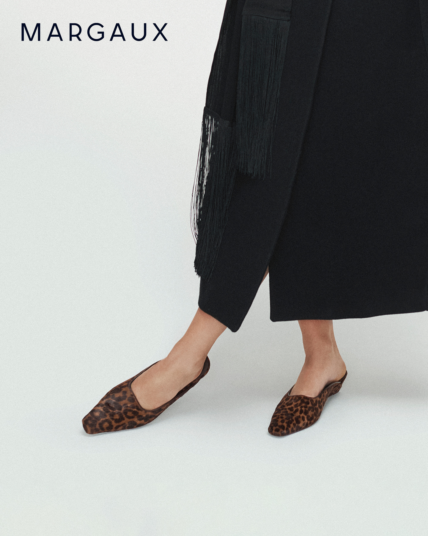 A close-up shot focuses on a person's feet wearing leopard-print slip-on shoes. The shoes have a pointed square toe and a low heel. The person is wearing a black garment with fringe details on the side. The brand name 'MARGAUX' is displayed in the upper left corner of the image. The text overlay highlights the soft Italian haircalf material, the squared toe, and the low heel of the shoes.