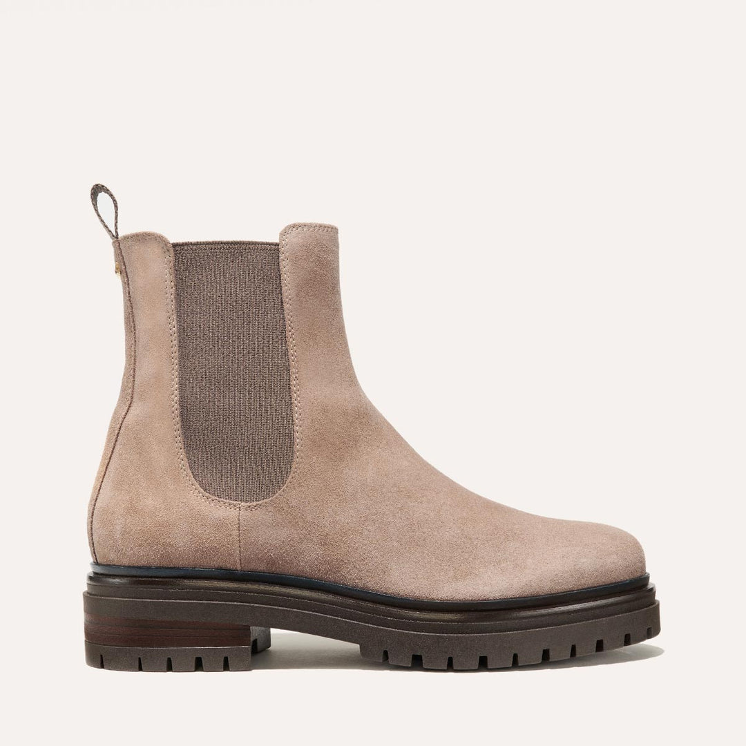A full-body studio shot of a single taupe suede Chelsea boot with a thick lug sole. The boot is shown from the side against a plain, light grey background. The copy is overlaid on the image, highlighting the comfort and style of the boots, emphasizing padded insoles and luxe materials.