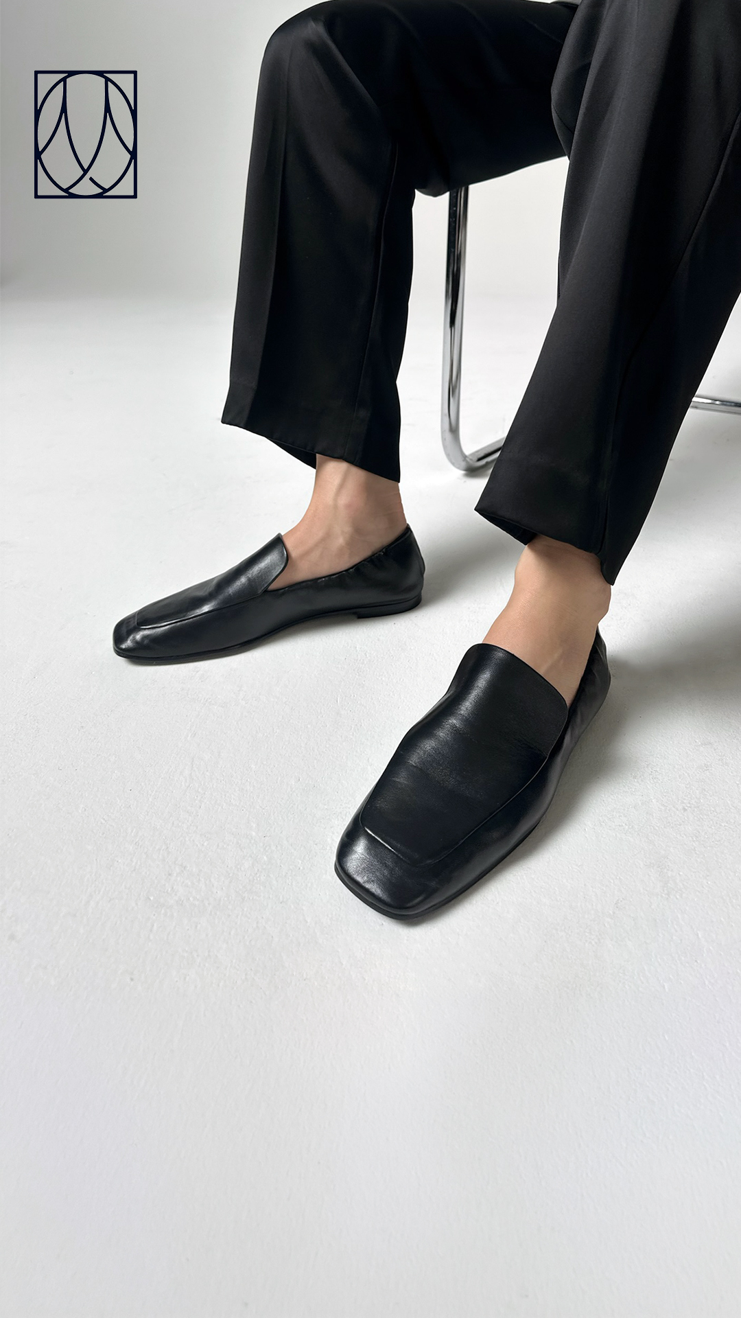 A close-up shot focuses on a person's feet wearing black, square-toed loafers. The person is wearing loose-fitting black pants, and their legs are crossed as they sit. The background is a plain white studio setting. The text overlay reads: "Our extra comfortable loafer is designed with perfectly ruched sides to fit like a glove."