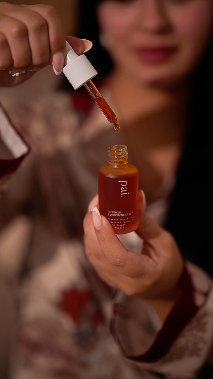 A close-up shot shows a woman's hands holding a small, amber-colored glass bottle of Pai Rosehip Bioregenerate facial oil. She is using the dropper to dispense a drop of the oil. The woman's face is softly blurred in the background, and she is wearing a robe with a floral pattern. Text overlays the image, stating 'Sensitive skin deserves skincare that’s both gentle and effective. 🧡 These two Pai essentials have become part of my evening routine, leaving my skin feeling clean, nourished, and naturally radiant.🧡 Now available exclusively at Ulta Beauty UAE online and in selected Ulta Beauty UAE stores. 🧡'.