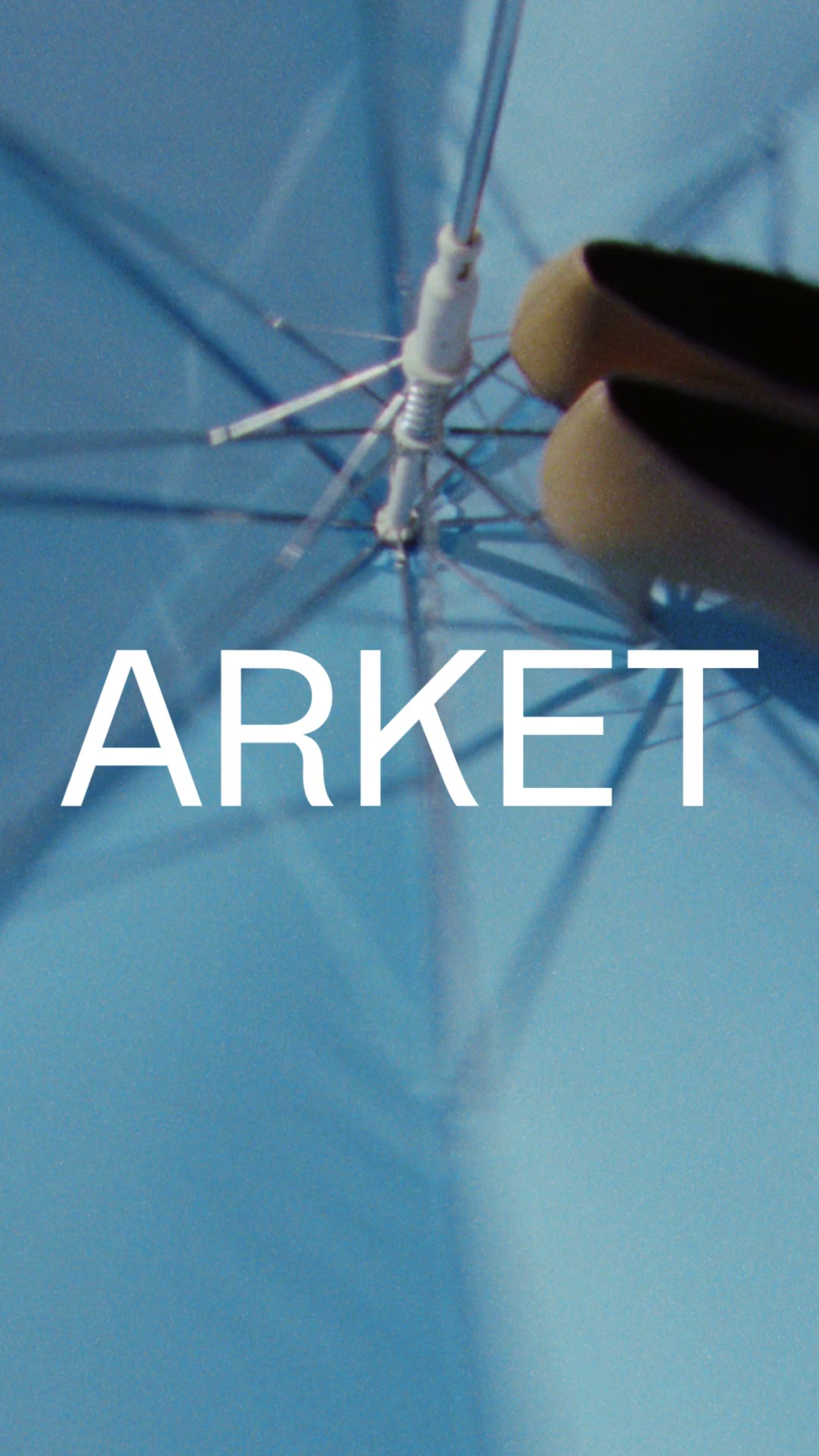 The creative is a vertical image with a light blue background, resembling the inside of an open umbrella. The brand name "ARKET" is displayed in large, white, sans-serif capital letters across the center of the image. Above the text, the metal structure of the umbrella and the tips of two brown shoes are visible, suggesting a fashion context. The overall aesthetic is minimalist and clean, with the text being the primary focus.
