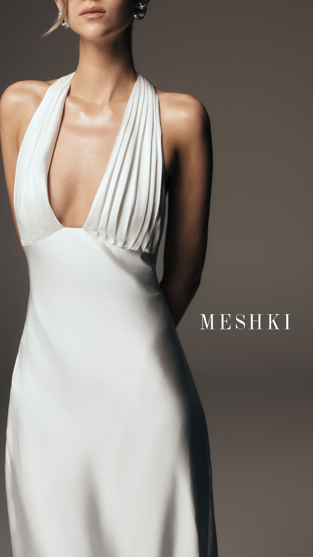 A close-up shot of a woman wearing a white halter dress with a plunging neckline and intricate pleating. The dress is made of a satin-like material that drapes elegantly. The woman is shown from the chest up, with her hands clasped behind her back. The brand name "MESHki" is displayed in white serif font on the right side of the image. The text overlay reads: "Designed for the moment you say yes to everything. From engagement to "I do" and beyond, MESHKI Bridal takes care of the details so you can be present for it all."