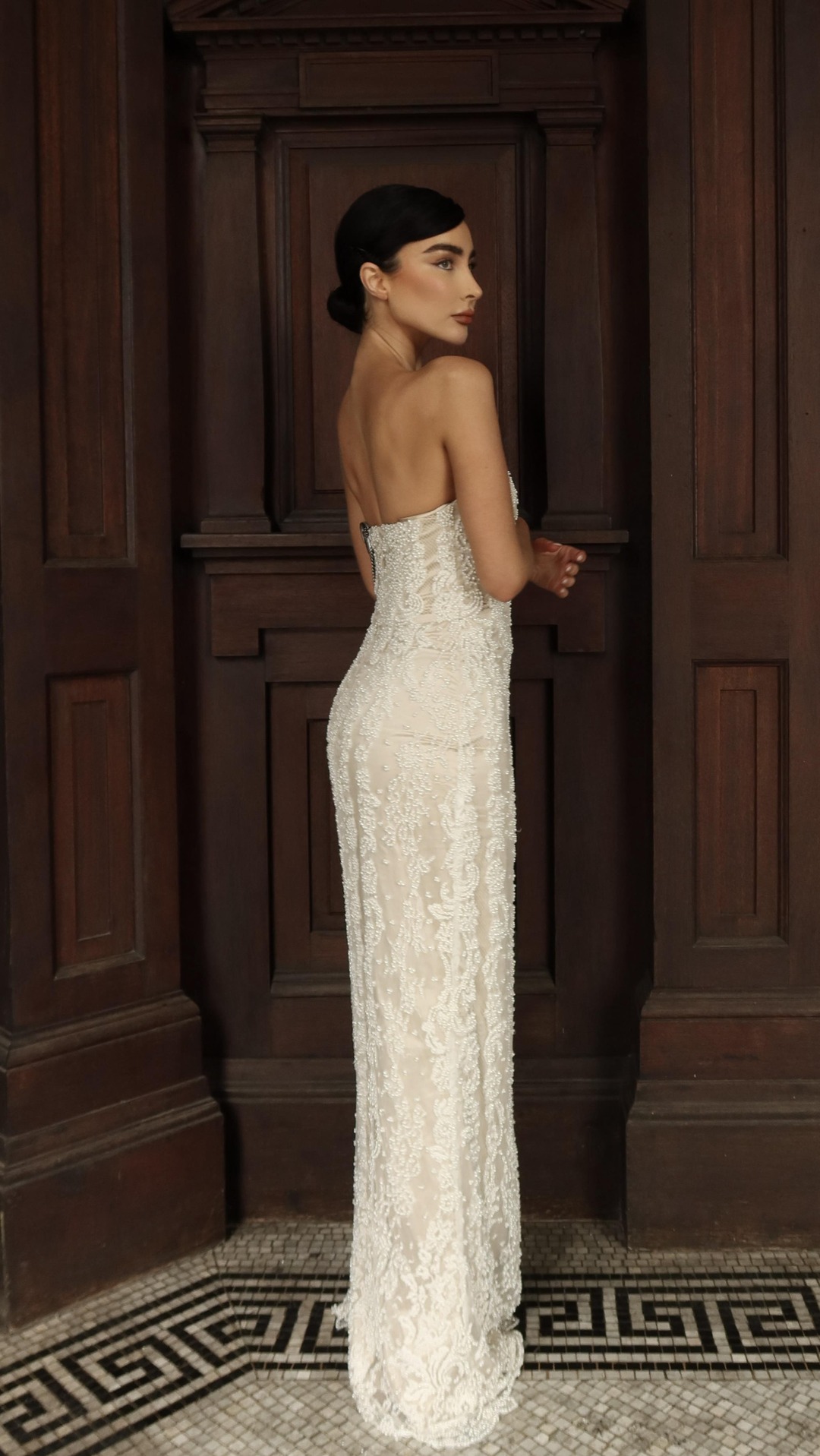 A woman stands in profile, wearing a long, strapless, white, beaded gown. She is positioned in front of a dark, ornate wooden door and architectural details. The text "Hand beaded to perfection — our EVA dress 🤍" is overlaid on the image. The creative focuses on the intricate detail and elegant design of the dress, presented in a sophisticated setting.