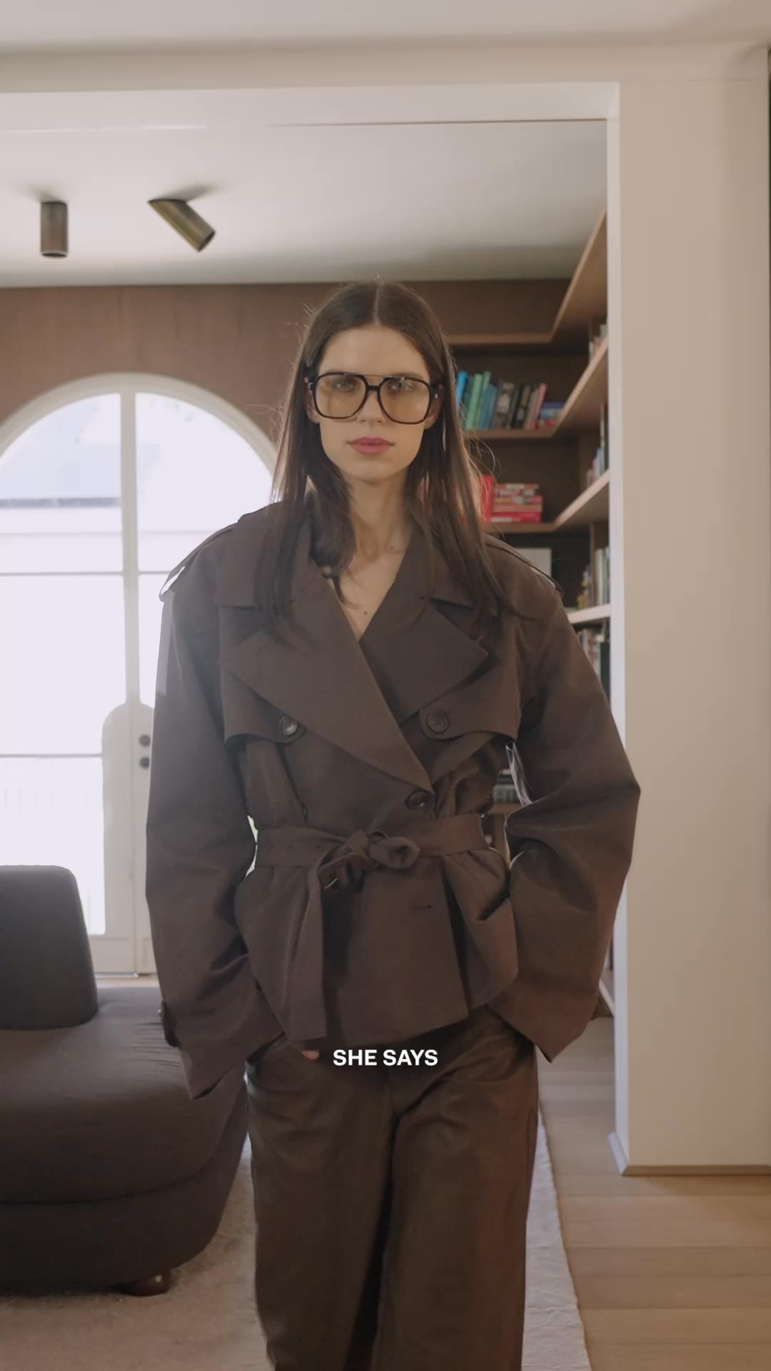 A woman wearing oversized glasses and a brown trench coat stands in a room with bookshelves in the background. She is looking directly at the camera. Text overlaid on the bottom of the image reads "SHE SAYS." The overall aesthetic is polished and sophisticated, suggesting a focus on professional or elevated fashion.