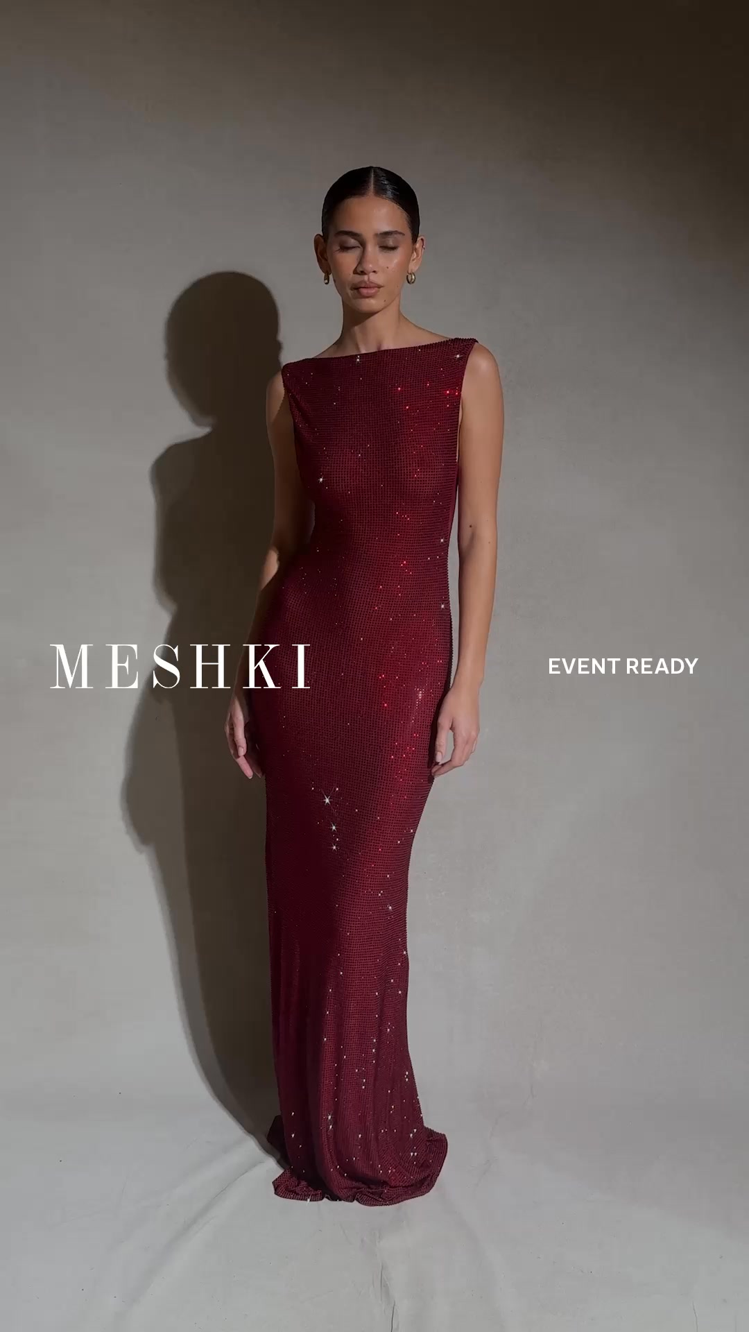A woman stands against a neutral grey background, wearing a long, form-fitting, deep red sequined dress. The dress has a boat neckline and is sleeveless. Her eyes are closed, and her head is slightly tilted. The brand name "MESHki" is overlaid in large, white serif font on the left side of the image, and the text "EVENT READY" is overlaid in smaller, white sans-serif font on the right side. The overall impression is elegant and sophisticated, suitable for formal occasions.