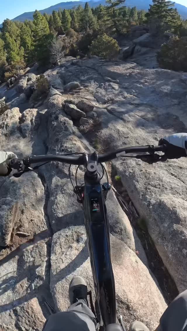 The video is a first-person point-of-view shot of someone riding an electric mountain bike over a rocky, outdoor trail. The rider's hands are on the handlebars, and their feet are on the pedals. The bike's display screen is visible, showing a speed of 3.4. Text overlays the video, stating, "eMTBs have never really been my thing, but the @aventon Current ADV caught my attention enough to give it a shot." The surrounding environment consists of trees and mountains under a clear blue sky.