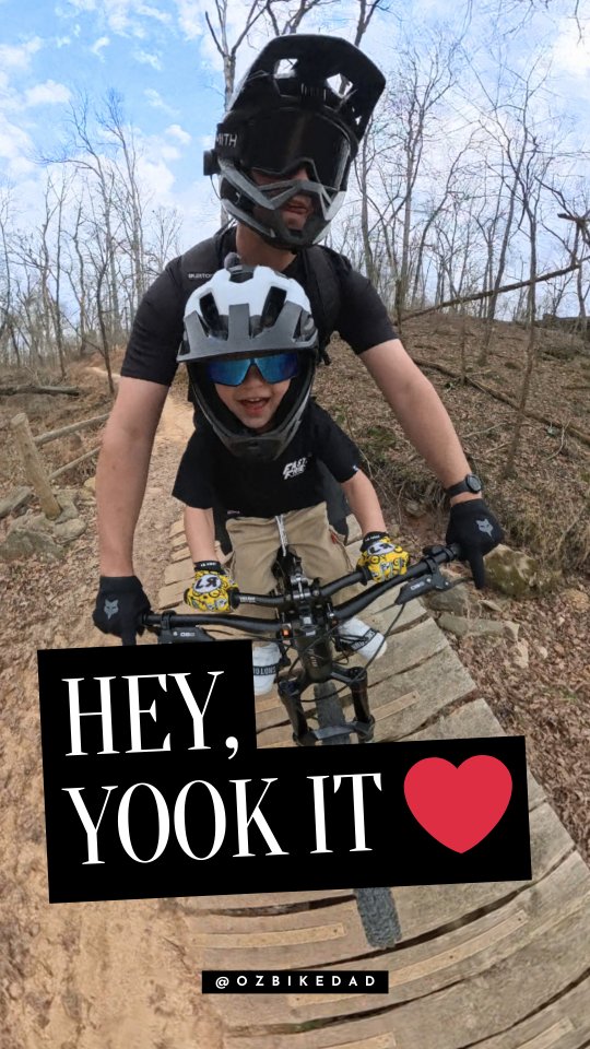 A first-person perspective shot of a father and child on a mountain bike trail. The father is wearing a full-face helmet and goggles, with the child seated in front of him on the handlebars, also wearing a helmet and sunglasses. Large white text on a black background reads "HEY, YOOK IT" with a red heart icon. The username "@OZBIKEDAD" is visible at the bottom of the image.