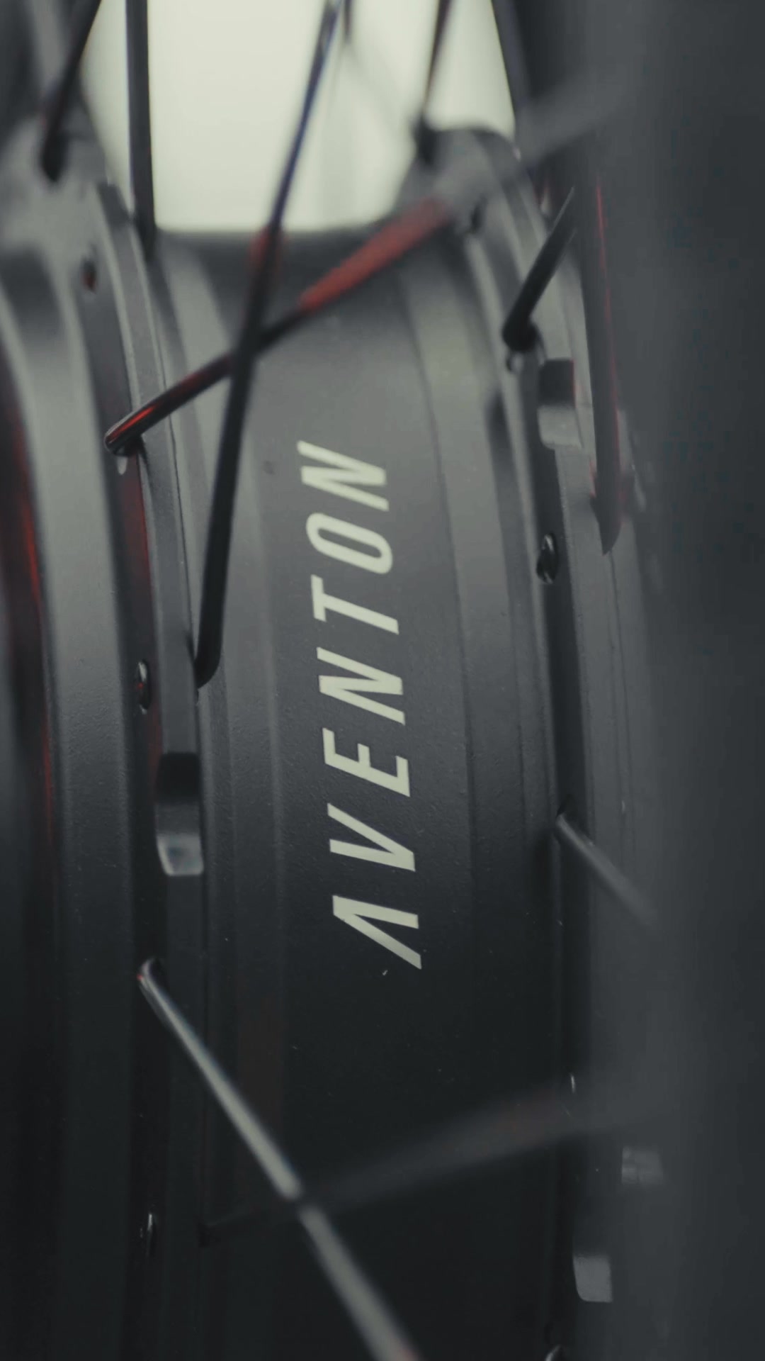 A close-up, shallow depth of field shot focuses on the hub of a bicycle wheel, specifically the black casing with the white Aventon logo printed vertically. The spokes of the wheel are visible, some in sharp focus and others blurred. The background is out of focus, with hints of red and grey tones. The text on the creative reads: "Our focus is your comfort with the Pace 5 REC. Stress-free at every touchpoint, the new Pace makes riding easier than ever before."