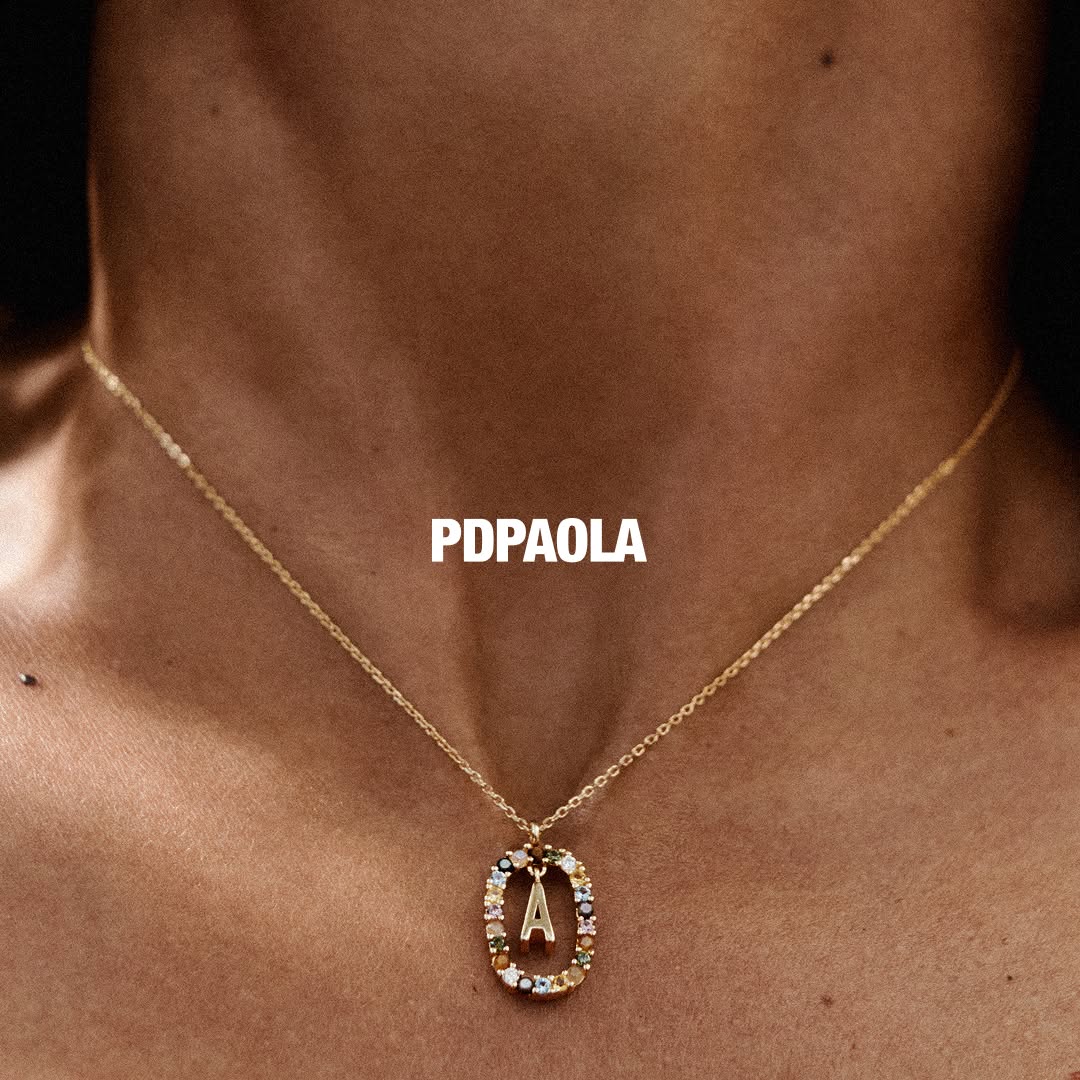 A close-up shot of a person's neck and collarbone, wearing a delicate gold necklace with a pendant. The pendant is an oval frame encrusted with colorful gemstones, and inside it, a gold letter 'A' is visible. The brand name 'PDPAOLA' is displayed in bold white text across the upper chest area. The copy 'A letter that says it all. Discover more.' is overlaid on the image.