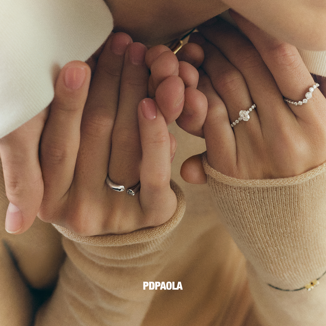 A close-up shot focuses on two pairs of hands intertwined, adorned with delicate rings. The hands are gently clasped, suggesting intimacy and connection. The text "PDPaOLA" is visible in the lower right corner. The overall aesthetic is soft and romantic, with a focus on the jewelry as symbols of affection.