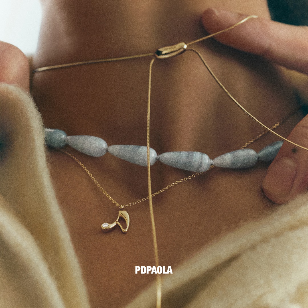 A close-up shot focuses on a person's neck, adorned with two delicate gold necklaces. The upper necklace features a strand of light blue, elongated beads, while the lower one showcases a small, elegant gold pendant with a single diamond. Hands are visible adjusting the chains, adding a touch of personal interaction. The brand name "PDPaola" is subtly displayed in white text at the bottom center of the image.