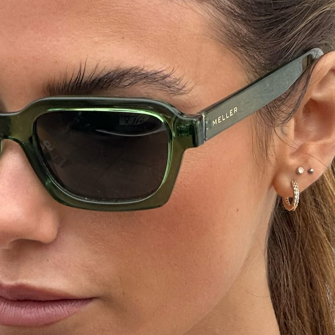 A close-up shot focuses on the right side of a woman's face, highlighting her wearing stylish green sunglasses with the brand name 'MELLER' subtly embossed on the temple. She also wears small hoop earrings and a stud earring. The lighting is soft, emphasizing the texture of her skin and the sleek design of the sunglasses. The image is a product shot with text overlay, showcasing the accessory in a fashionable context.