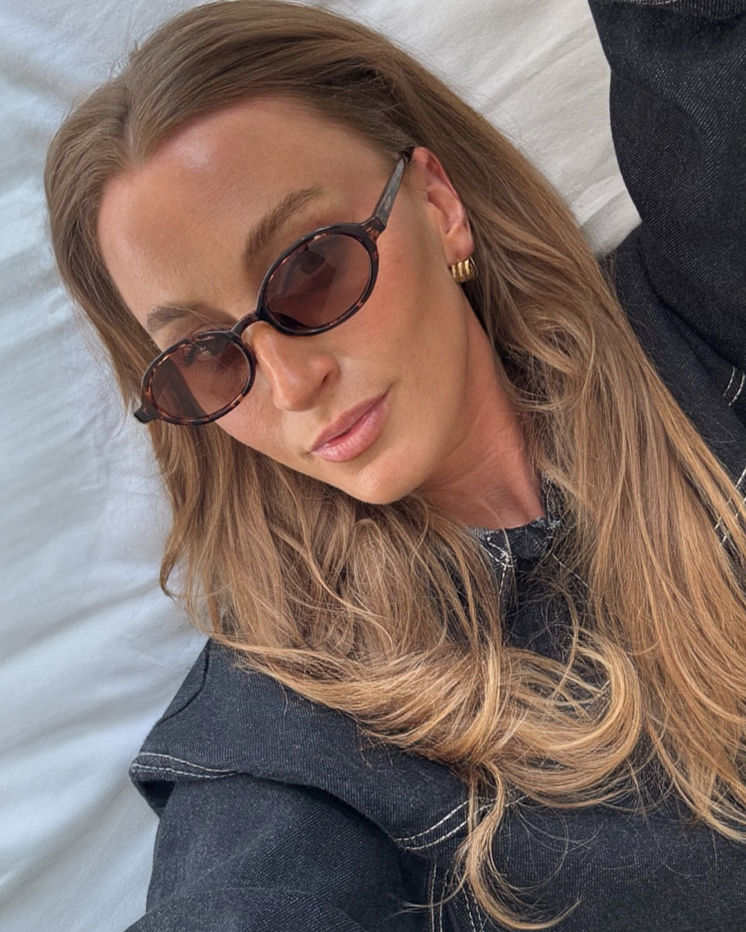 A close-up, overhead shot shows a woman with long, wavy blonde hair wearing oval-shaped, tortoiseshell sunglasses and a dark denim jacket. She is lying down on a white surface, looking slightly away from the camera. The text "Your new favorite sunglasses are here." is overlaid in a simple, white font on the right side of the image. The overall aesthetic is casual and stylish, suggesting a focus on fashion and personal style.