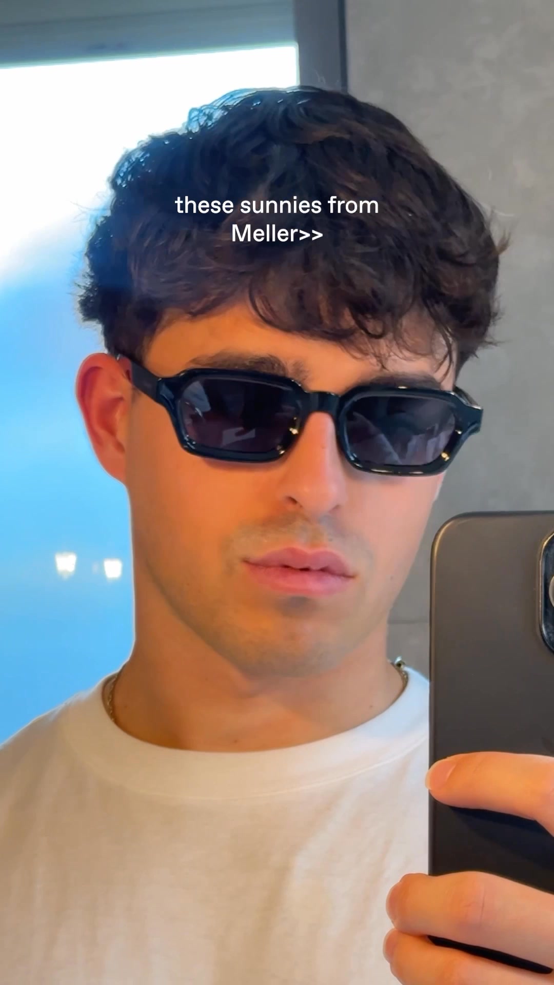 A close-up selfie of a young man wearing black, rectangular sunglasses. He is looking directly at the camera, with his mouth slightly open. White text overlaid on the image reads "these sunnies from Meller>>". The background is blurred, with hints of blue and grey. The man is wearing a white t-shirt and a thin chain necklace.