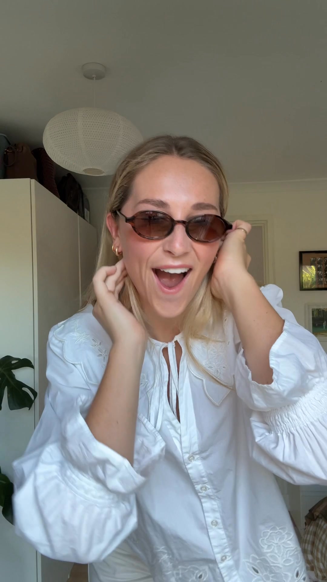 A young woman with blonde hair is wearing sunglasses and a white collared shirt. She is looking at the camera with an excited expression, touching her sunglasses and hair. The text overlay reads "POV: You finally found the perfect sunglasses." The background shows a closet and a lamp.