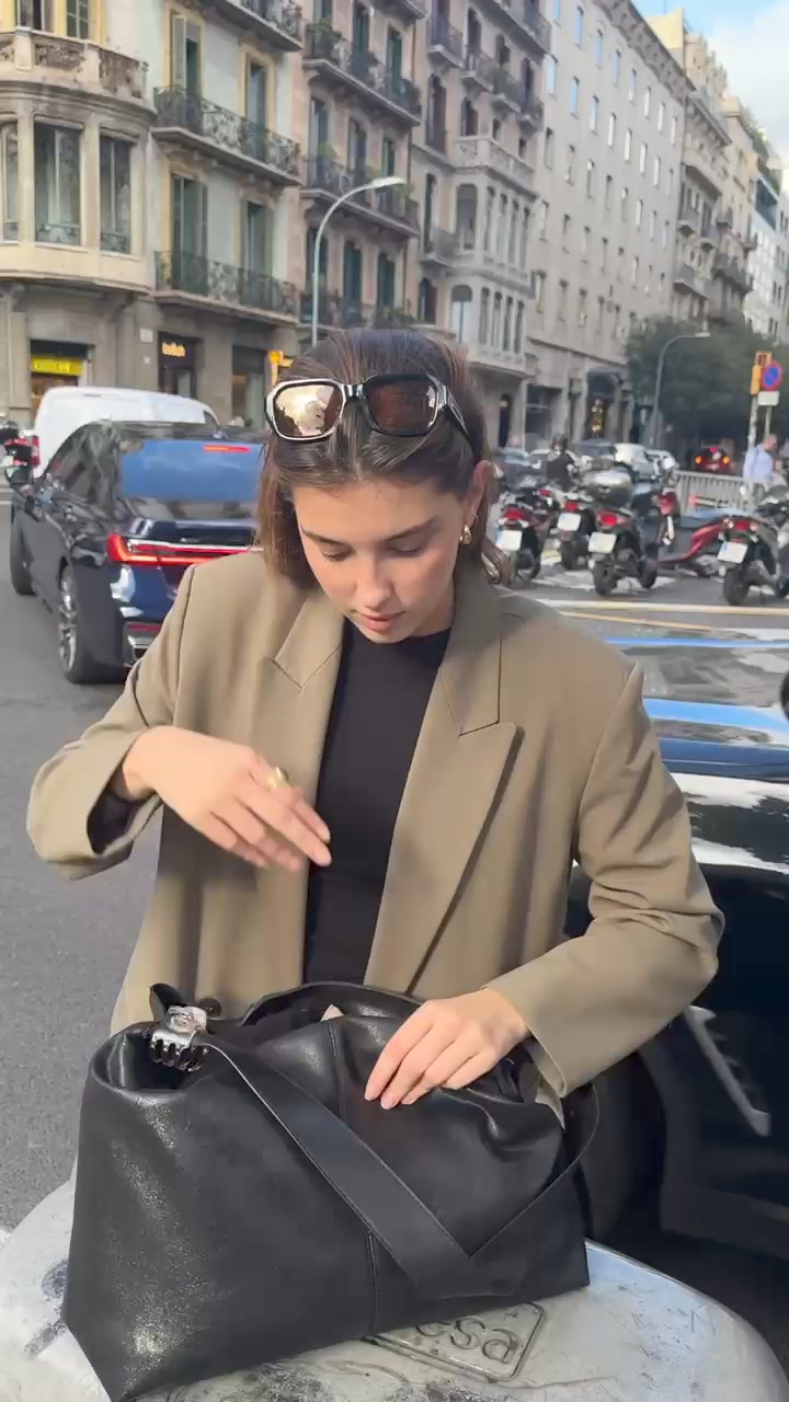 A young woman wearing sunglasses on her head and a beige blazer over a black top is shown in an urban setting. She is looking down and reaching into a large black handbag. Text overlays the image, stating "Your new favorite sunglasses are here." The background features buildings and parked motorcycles.