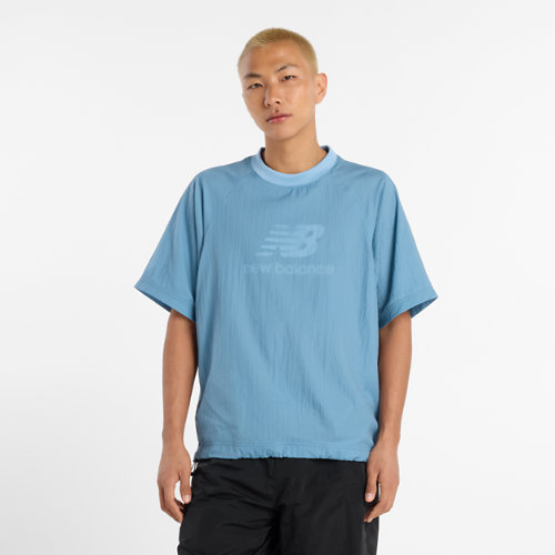 A young man with short, bleached blonde hair stands against a plain white background, wearing a light blue New Balance t-shirt and black shorts. The t-shirt has the New Balance logo embossed in a slightly darker shade of blue on the chest. The text "Unsere vielfältige Auswahl an Produkten - für jeden Lifestyle" is overlaid on the image in a simple, sans-serif font. The overall presentation is clean and focuses on the apparel.