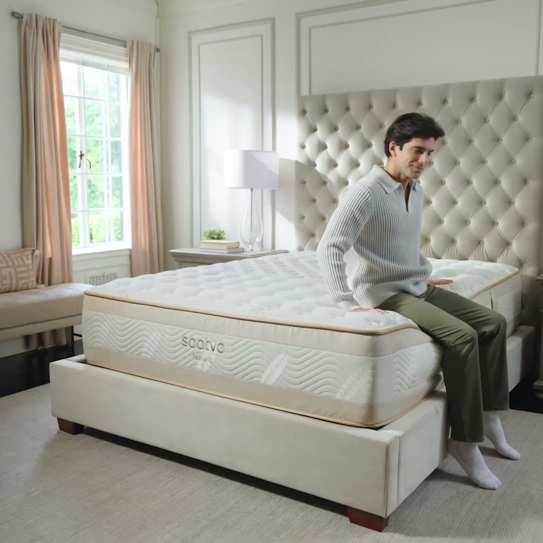 A man is sitting on the edge of a Saatva mattress in a well-decorated bedroom. He is wearing a light-colored sweater and dark pants, and he is looking down with a slight smile. The mattress is white with gold trim and has the Saatva logo visible. The room has a large tufted headboard, a window with light pink curtains, and a bedside table with a lamp. The on-screen text reads: "How you wake up shapes the rest of your day."