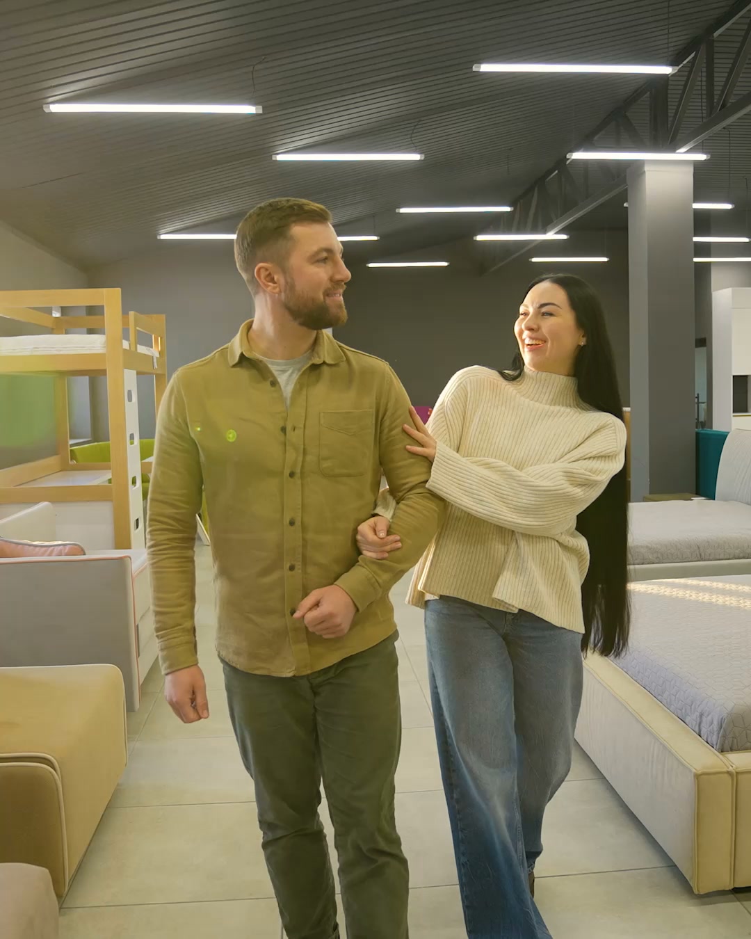 A man and a woman walk arm-in-arm through a brightly lit mattress store, smiling at each other. The man wears a khaki button-down shirt and olive pants, while the woman wears a cream-colored sweater and blue jeans. The background shows various beds and bunk beds. Text overlays the image, explaining that Saatva offers luxury mattresses without the traditional retail markup by selling directly to consumers.
