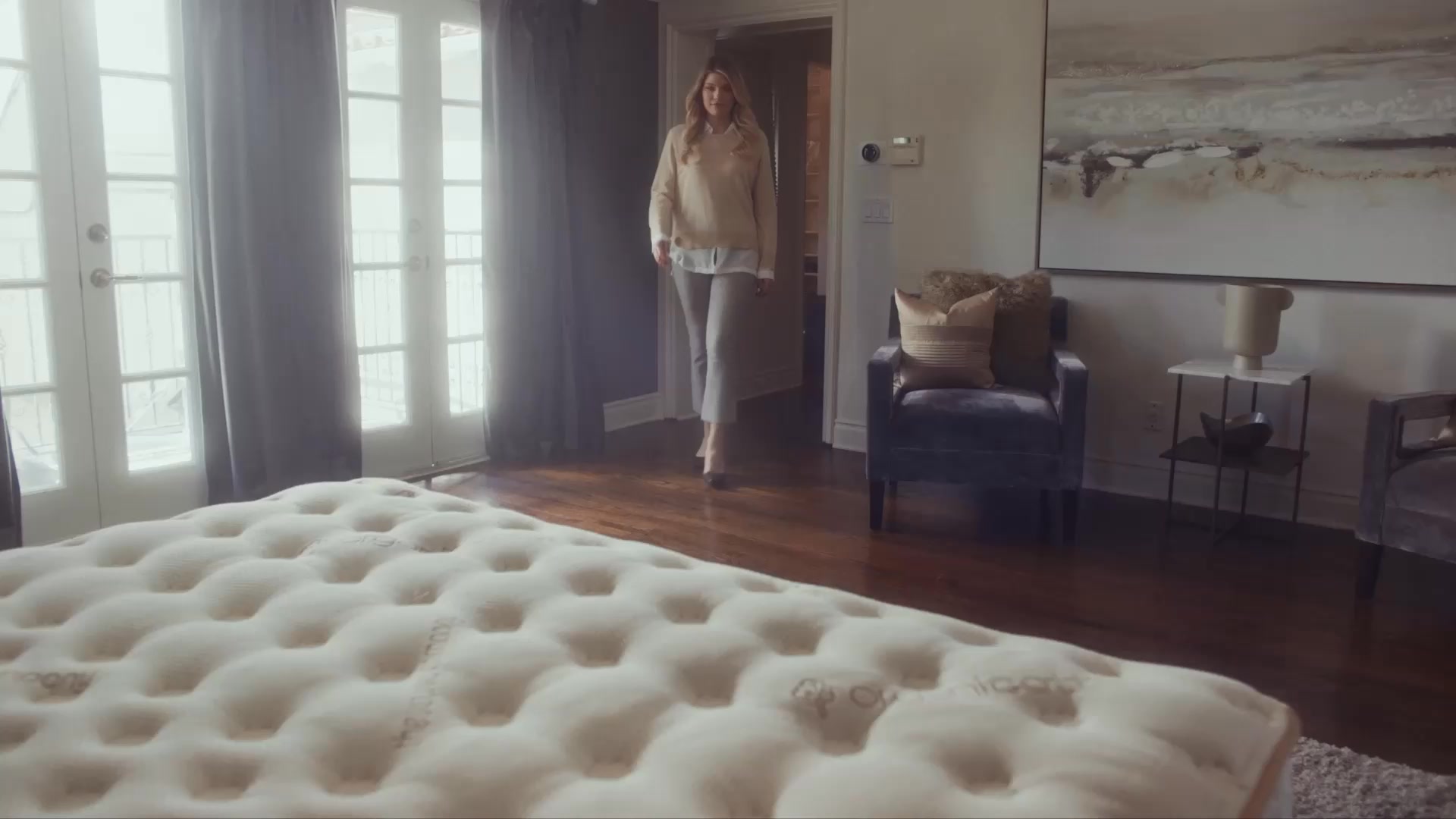 A woman in casual professional attire walks into a well-decorated bedroom. The room features a large tufted ottoman in the foreground, a plush armchair, and a large abstract painting on the wall. The text "Prioritize Your Sleep. Start with Saatva." is overlaid on the image. The overall impression is one of comfort and sophisticated home decor.