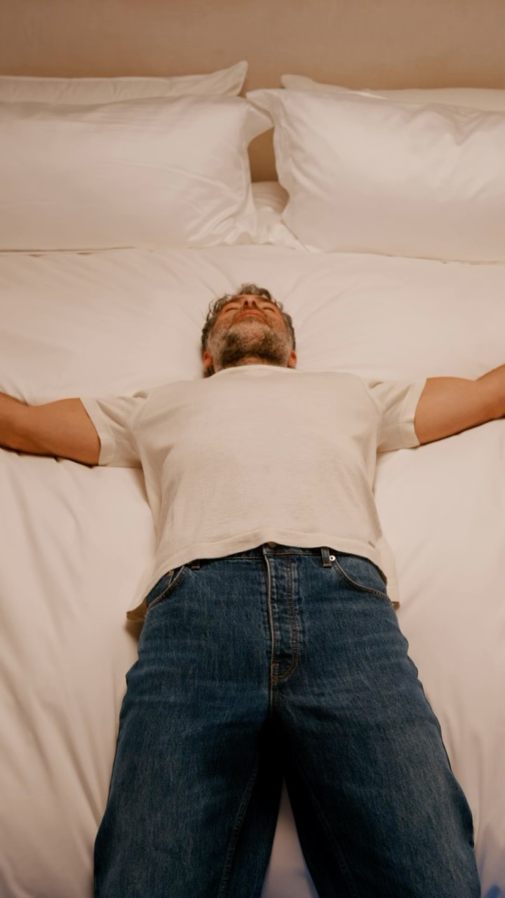 A man lies on his back in a bed with white sheets and pillows, arms spread wide, eyes closed, and a relaxed expression. He is wearing a white t-shirt and blue jeans. White text overlaid on the image reads "Resting up for Fashion Week with @saatva #saatvapartner".