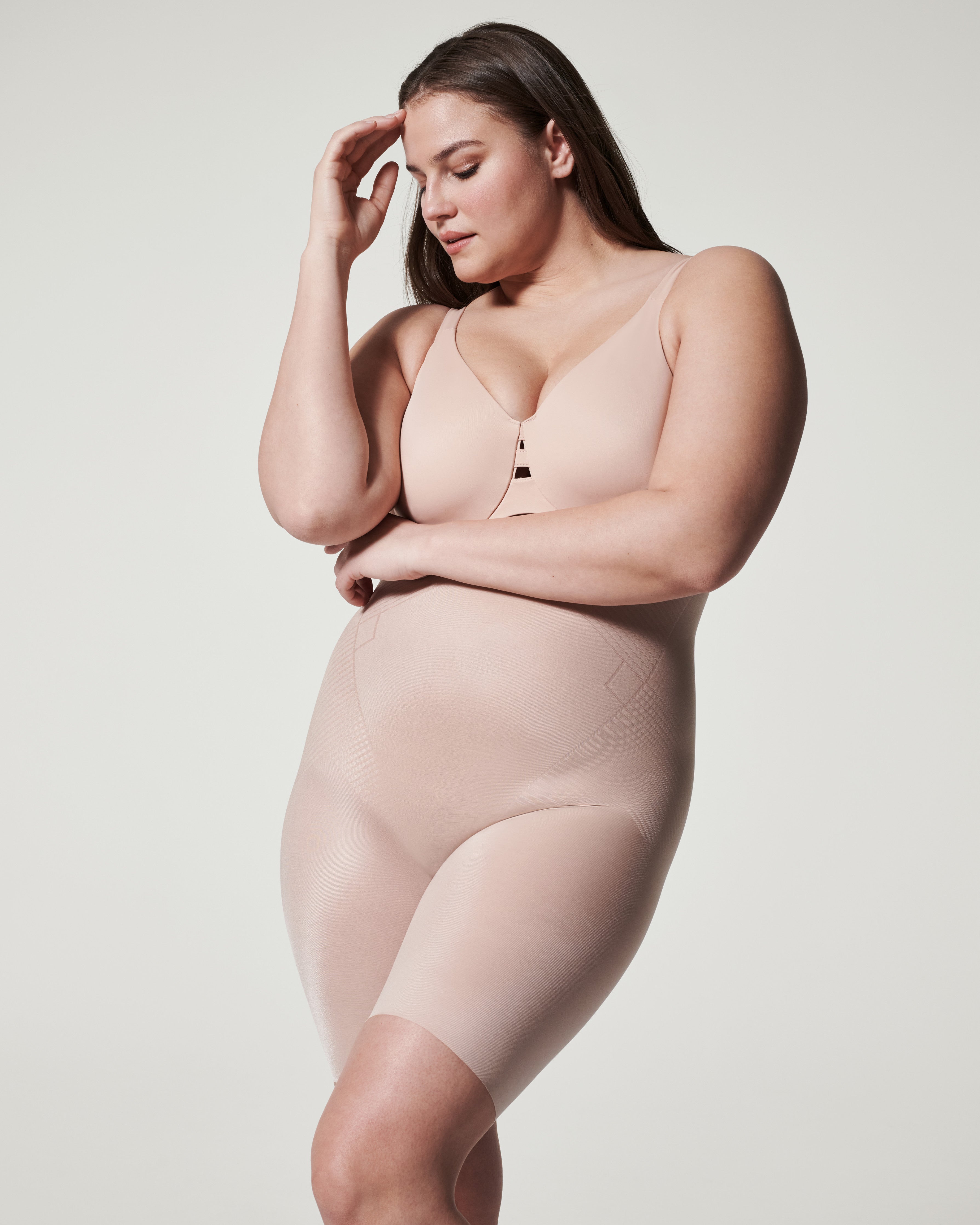 A plus-size model poses in a neutral-toned bodysuit against a plain, light grey background. She is looking down with her hand on her forehead, conveying a sense of contemplation or perhaps relief. The bodysuit appears to be shapewear, with subtle textured panels on the sides of the torso and thighs. The text "Meet the newest must-haves!" is overlaid on the image in a simple, sans-serif font.