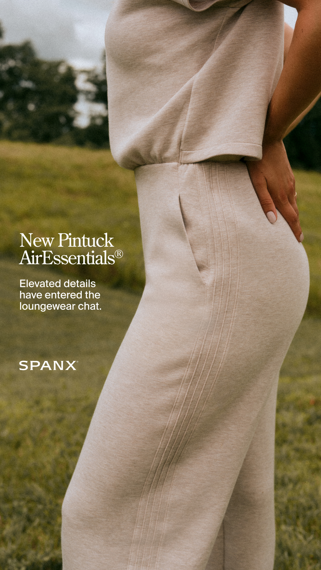 A close-up, side-profile shot of a person wearing a light beige loungewear set outdoors. The text "New Pintuck AirEssentials®" is overlaid on the left side of the image, followed by the descriptive text "Elevated details have entered the loungewear chat." Below this, the "SPANX" logo is displayed. The image focuses on the texture and details of the clothing, such as the pintuck stitching on the pants and the soft fabric.