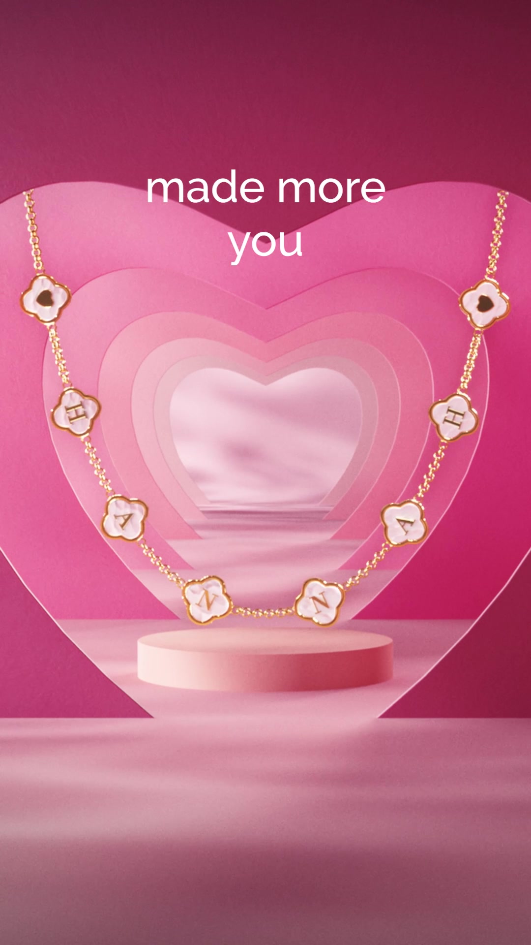 A gold necklace with white clover-shaped charms, each containing a letter, is displayed against a pink, layered heart-shaped backdrop. The letters spell out "HANNA". White text overlays the image, reading "made more you". The necklace is presented on a small pink pedestal.