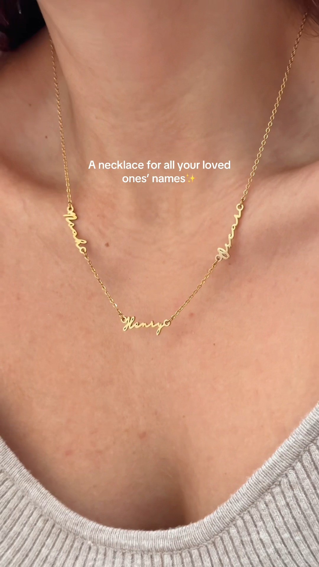 A close-up shot shows a gold necklace with three names written in cursive hanging from it. The names are 'Noah', 'Henry', and 'Oscar'. Text overlaid on the image reads "A necklace for all your loved ones' names ✨". The background is the skin of a person's neck and chest, wearing a grey ribbed shirt. The creative focuses on the sentimental value of personalized jewelry.