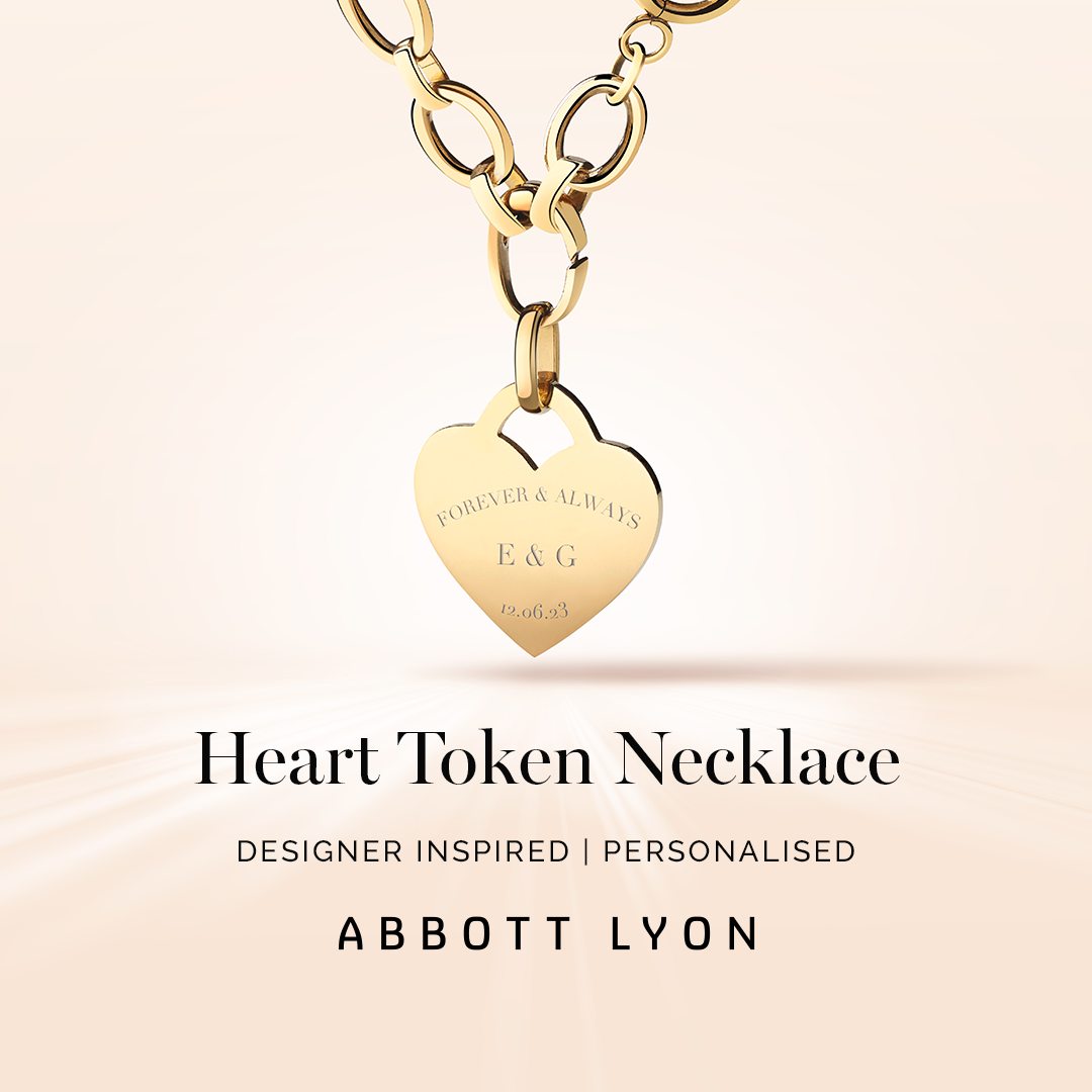 A close-up shot of a gold heart-shaped pendant necklace with the words "FOREVER & ALWAYS", "E & G", and "12.06.23" engraved on it. The necklace hangs from a chunky gold chain. Below the necklace, the text "Heart Token Necklace" is displayed prominently, followed by "DESIGNER INSPIRED | PERSONALISED" and the brand name "ABBOTT LYON". The background is a soft, gradient of peach and cream with subtle light rays emanating from the bottom.