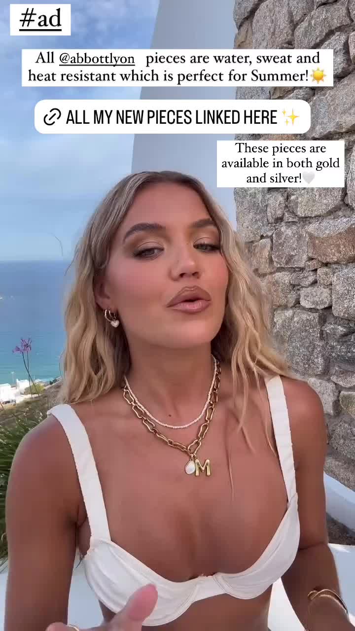 A woman with blonde hair, wearing a white bikini top and necklaces, is shown outdoors with a body of water and stone wall in the background. Text overlays on the image read: "#ad", "All @abbottlyon pieces are water, sweat and heat resistant which is perfect for Summer! ☀️", and "ALL MY NEW PIECES LINKED HERE ✨". Another text box states, "These pieces are available in both gold and silver!"